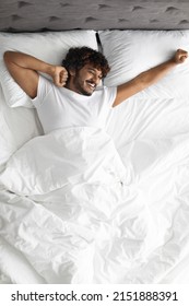 Happy curly hindu guy in white t-shirt stretching body while laying in comfortable bed, looking at copy space and smiling, enjoying new day, feeling fully rested, top view, vertical shot