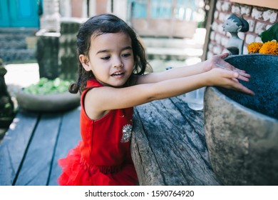 Happy Curly Little Girl In Red Dress Washing Hands In Stone Sink Standing On Outside Terrace In Ethnic Style Looking Down