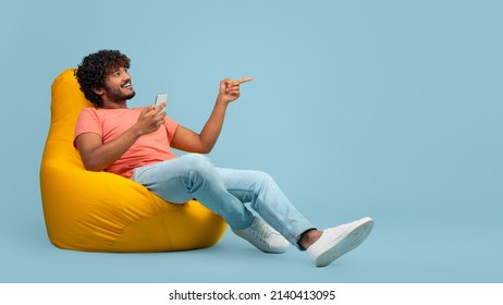Happy curly millennial indian man sitting at bean bag with brand new smartphone and pointing at copy space over blue studio background, showing nice offer and smiling, panorama