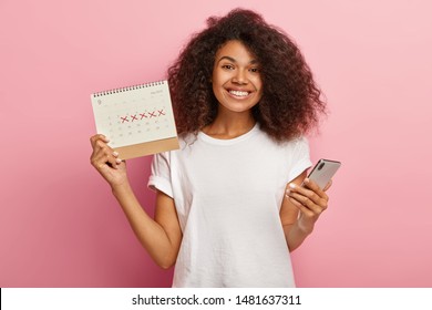 Happy Curly Woman Holds Periods Calendar, Checks Menstruation Days On Mobile Phone Application, Cares About Women Health, Dressed Casually, Isolated On Pink Studio Wall. Pms And Wellness Concept