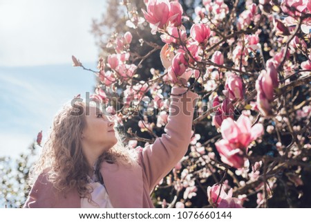happy curly woman in a pink coat enjoying a fragrant pink magnolia in a park