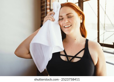 Happy curvy woman with red hair, plus-size, wiping sweat with a towel. Plus-size woman in workout attire, smiling. Red-haired woman, plus-size, cooling down after exercise. Wellness routine. - Powered by Shutterstock