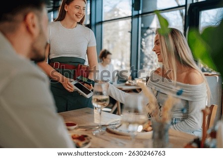 Happy customer paying bill using contactless payment with smartphone in a restaurant, waitress holding pos terminal