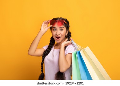 Happy Customer. Portrait Of Excited Emotional Young Indian Woman Wearing And Touching Sunglasses Holding Colorful Shopping Bags With Gifts, Amazed Lady Looking At Camera, Yellow Studio Background