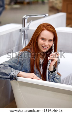 happy customer sitting inside of new bathroom, playing fool. young redhead female in denim jacket have fun, talk on handle
