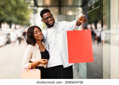 Happy Customers Concept. Portrait of smiling African American couple walking near shopping centre, looking at showcase, guy pointing finger at mall window. Seasonal Sales And Discount, Special Offer
