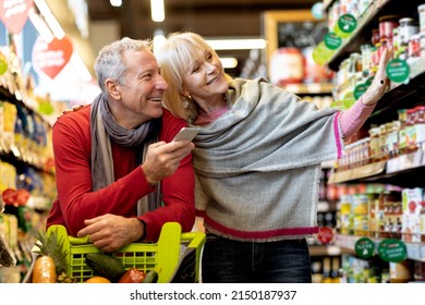 Happy customers senior couple doing grocery, shopping and using smartphone, walking with cart in supermarket. Man and woman using shopping application on phone, buying food in supermarket Foto Stock
