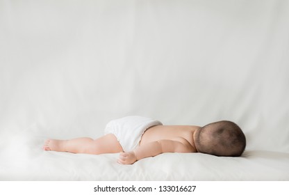 Happy Cute 5 Month Old Asian Baby Boy With Short Black Hair Wearing A White Cloth Nappy And Sleeping On His Front On A White Bed