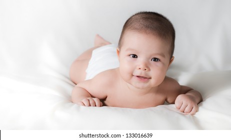 Happy Cute 5 Month Old Asian Baby Boy With Short Black Hair Wearing A White Cloth Nappy And Lying On His Front On A White Bed