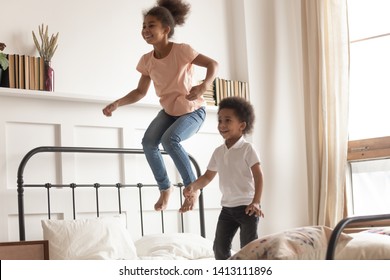 Happy Cute Active Little African American Kids Boy And Girl Jumping On Bed Laughing Together, Two Funny Small Energetic Mixed Race Children Brother With Sister Having Fun Play Active Game In Bedroom