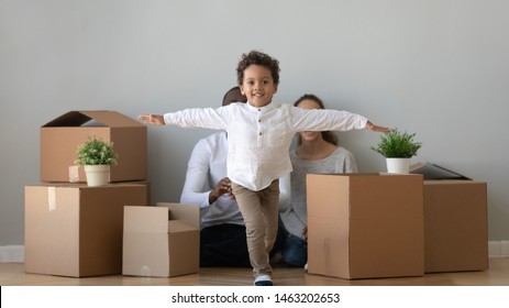 Happy Cute African Child Boy Running In New Apartment Looking At Camera On Moving Day, Excited Little Kid Son Exploring House With Parents Boxes On Background, Children And Family Mortgage Relocation