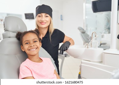 Happy Cute African Girl Sitting In Dental Chair, Smiling And Looking At Camera After Oral Hygiene By Professional Beautiful Dentist In Black Medical Uniform. Concept Of Treatment And Happiness.