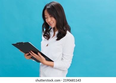 Happy Cute Asian Businesswoman In Classic Office Dress Code Holds Folder Tablet Read Candidate's Resume Posing Isolated On Over Blue Studio Background. Cool Business Offer. Job Interview Concept