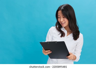 Happy Cute Asian Businesswoman In Classic Office Dress Code Holds Folder Tablet Read Candidate's Resume Posing Isolated On Over Blue Studio Background. Cool Business Offer. Job Interview Concept