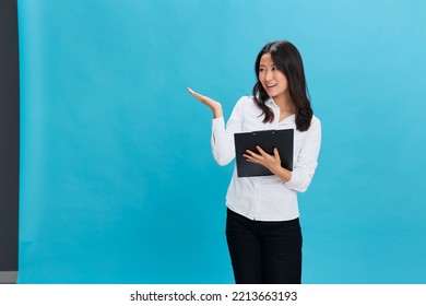 Happy Cute Asian Businesswoman In Classic Office Dress Code Holds Folder Tablet Point Hand Up At Cope Space Posing Isolated On Over Blue Studio Background. Cool Business Offer. Job Interview Concept