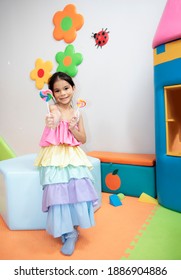 Happy Cute Asian Child Girl Wearing Colourful Dress Eating Candy Lollipop. Adorable Kid Girl With Colorful Lollipop. Portrait Little Girl With A Lollipop Stick In Birthday Party Toy At Cafetoyer.