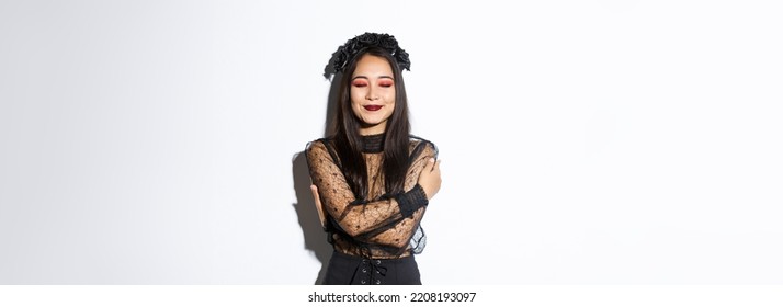 Happy Cute Asian Woman Hugging Herself And Smiling Dreamy, Wearing Halloween Costume Of Witch Or Widow, Standing Over White Background