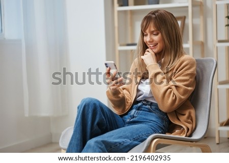 Happy cute blonde student lady in warm sweater chatting with boyfriend use phone sitting in armchair at home interior. Pause from work, take a break, social media in free time concept. Copy space