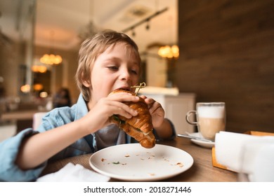 Happy cute boy eating fresh croissant, sitting at the table in city cafe, with paper cup of cocoa. High quality photo - Powered by Shutterstock