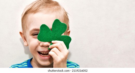 Happy cute boy hiding one eye behind clover leaf on light background. Happy St. Patrick's Day. Portrait of a child. Concept holiday. Banner with copy space