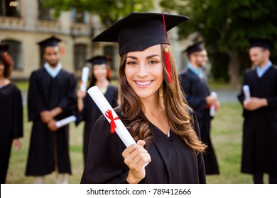 Happy cute brunette caucasian grad girl is smiling, blurred class mates are behind. She is in a black mortar board, with red tassel, in gown, with nice brown curly hair, diploma in hand - Powered by Shutterstock