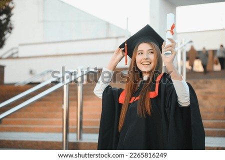 Happy cute brunette caucasian grad girl is smiling. She is in a black mortar board, with red tassel, in gown, with nice brown curly hair, diploma in hand