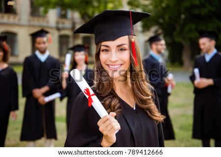 Happy cute brunette caucasian grad girl is smiling, blurred class mates are behind. She is in a black mortar board, with red tassel, in gown, with nice brown curly hair, diploma in hand
