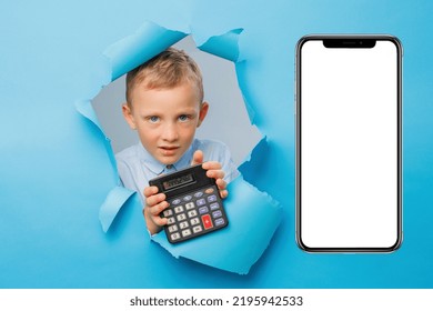 Happy Cute Businessboy Is Having Fun And Holding Calculator In Hand On Blue Background Wall, Climbs Through A Hole In The Paper Near Huge Mobile Phone. Bright And Funny Emotions Of The Boy