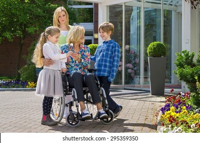 Happy cute caring grandchildren with concerned mother visiting youthful looking grandmother in wheelchair.