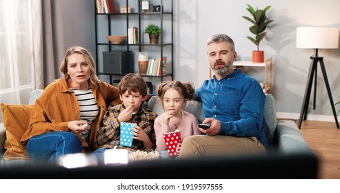 Happy Cute Caucasian Family Resting At Home Watching TV Spending Time Together With Popcorn And Shocked Scared Faces Mom And Dad With Small Kids Eating Popcorn Watching Horror Movie Closing Child Eyes