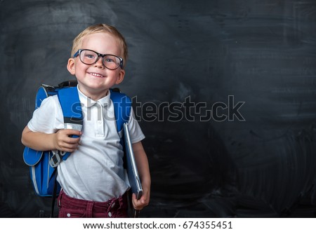 Happy cute clever boy in glasses with school bag and book in his hand.  Modern backpack. Child is ready to answer with a blackboard on a background. First time to school. Back to school. 