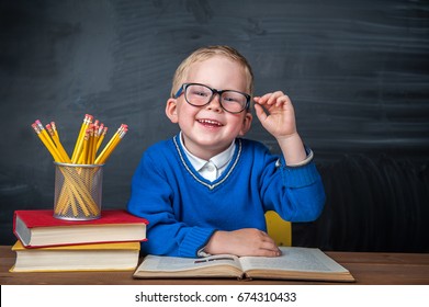 Happy cute clever boy is sitting at a desk in a glasses with raising hand. Child is ready to answer with a blackboard on a background. Ready for school. Back to school. Apple and books on desk - Shutterstock ID 674310433