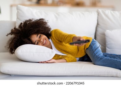 Happy Cute Curly African American School Girl Lying On Couch Alone, Holding Remote Controller, Black Kid Watching TV At Home, Switching Channels, Enjoying Nice Cartoon Or Show, Copy Space