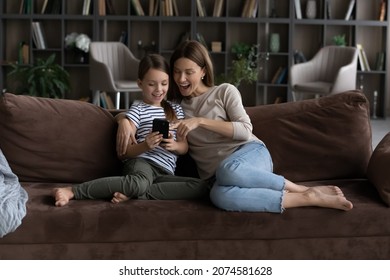 Happy Cute Daughter Kid With Mother Using Smartphone At Home, Sitting On Couch Together, Smiling Girl Child With Mom Looking At Phone Screen, Watching Funny Video, Browsing Apps, Taking Selfie