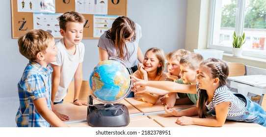 Happy cute enthusiastically pupils smiling around a globe in classroom with female teacher at school. Group of curious classmates at workplace studying globe in classroom. - Shutterstock ID 2182378951