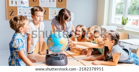 Happy cute enthusiastically pupils smiling around a globe in classroom with female teacher at school. Group of curious classmates at workplace studying globe in classroom.