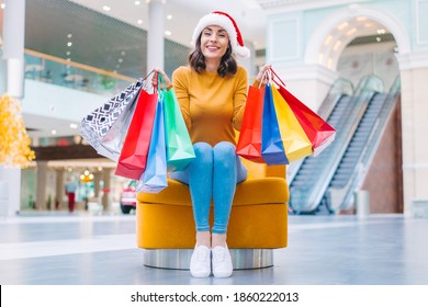 Happy Cute Girl In Santa Clause Hat Sitting On The Chair In The Mall With Bunches Of Colorful Shopping Bags In Hands. Christmas And New Year Shopping Concept.