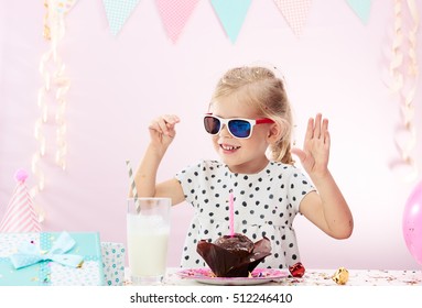 Happy Cute Girl In Sunglasses Celebrating Festive Event Indoors. Little Child Sitting At The Table With Milk Cocktail, Chocolate Cupcake, Birthday Gift. 