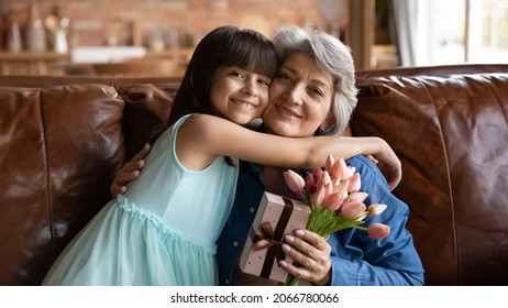 Happy Cute Granddaughter Girl Hugging Grandma On Sofa At Home Due To Birthday, 8 March Celebration. Grandmother Holding Gift Wrap, Flowers, Embracing Beloved Grandkid, Looking At Camera, Smiling