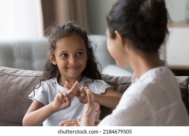 Happy cute Indian kid girl talking to mother, using hands, fingers, speaking sign languages, sitting on sofa at home. Female teacher, therapist teaching child with deafness to communicate - Shutterstock ID 2184679505