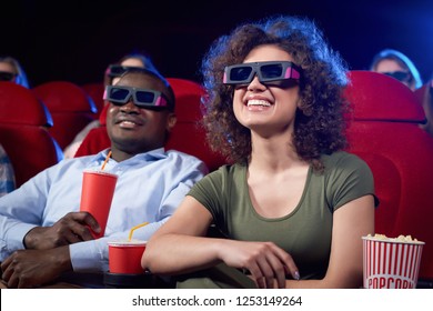 Happy cute international couple eating popcorn and laughing at funny comedy in cinema theater. Attractive girl and handsome afro man having romantic date and enjoying interesting movie.