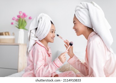 Happy Cute Korean Teen Girl And Millennial Female In Pink Pajamas And Towel Applying Powder To Face, Sitting On Bed In Bedroom Interior, Profile. Home Treatments, Makeup, Beauty Care And Relationships