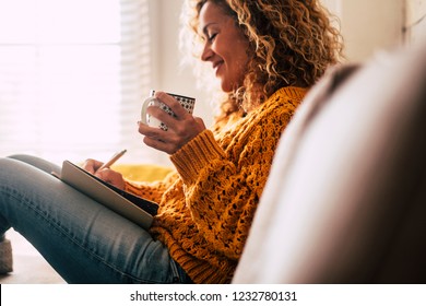 Happy cute lady at home write notes on a diary while drink a cup of tea and rest and relax taking a break. autumn colors and people enjoying home lifestyle writing messages or lists. Blonde curly