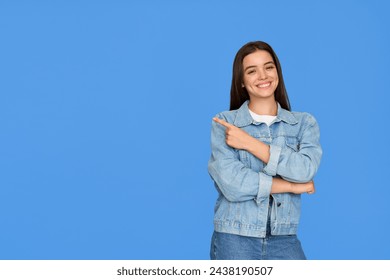 Happy cute Latin teenage student girl pointing aside standing on background. Smiling pretty hispanic teenager with brunette hair wearing denim jacket presenting ads standing isolated on blue. - Powered by Shutterstock