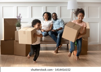 Happy Cute Little African Kids Holding Boxes Run Play In Living Room While Parents Relax On Moving Day, Black Family Renters Tenants With Children Celebrate Buy New Home Having Fun Renovate House