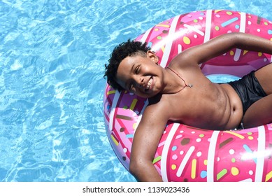 Happy Cute Little African-american Boy Lying On An Inflatable Donut Circle  In The Swimming Pool In The Hotel. Cool Summer Holidays For Children And Kids. Active Games On The Water.  Pink Donat. 