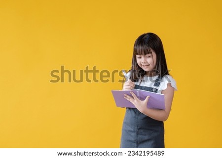 A happy and cute little Asian girl is writing something in her book, standing against an isolated yellow background. schoolgirl, student, children, elementary school student
