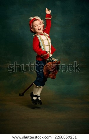 Happy cute little boy dressed up as medieval little prince and pageboy ride toy horse over dark vintage style background. Vintage fashion, emotions, theater art concept. Eras comparison