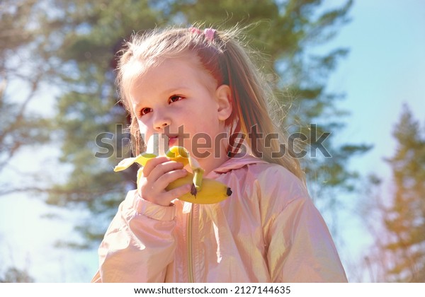 Happy cute little girl with banana fruit.\
Walking outdoors, healthy food eating\
concept.
