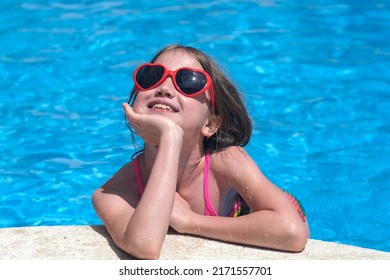 Happy Cute Little Girl In Heart Shaped Sunglasses Dreaming In Swimming Pool On Sunny Day. Childhood, Valentine's Day And Summer Vacation Concept. Smiling Child Having Fun Outdoors On Family Holidays